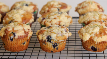 A batch of blueberry muffins topped with crumbly streusel sits on a cooling rack. The muffins are golden brown with visible blueberries peeking through the baked batter. The cooling rack is placed on a white surface.