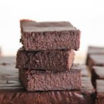 A stack of three chocolate brownies sits on top of a larger array of brownies, all cut into squares. The brownies have a rich, dense texture and a dark chocolate color. The background is out of focus, emphasizing the dessert.