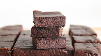 A stack of three chocolate brownies sits on top of a larger array of brownies, all cut into squares. The brownies have a rich, dense texture and a dark chocolate color. The background is out of focus, emphasizing the dessert.