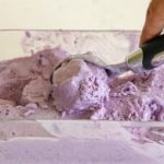 A hand is scooping purple ice cream from a clear rectangular container with a black-handled ice cream scoop. The ice cream has a smooth, creamy texture and is partially scooped out, revealing the container's bottom.