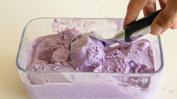 A hand is scooping purple ice cream from a clear rectangular container with a black-handled ice cream scoop. The ice cream has a smooth, creamy texture and is partially scooped out, revealing the container's bottom.