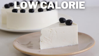 A slice of low-calorie cheesecake topped with blueberries placed on a beige plate. The remaining cheesecake, also garnished with blueberries, is visible in the background on a white serving dish. The words "LOW CALORIE" are prominently displayed at the top of the image.