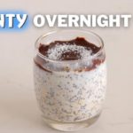 A glass of overnight oats topped with a layer of chocolate and sprinkled with coconut flakes on a white surface. Bold white text above the glass reads "Bounty Overnight Oats.