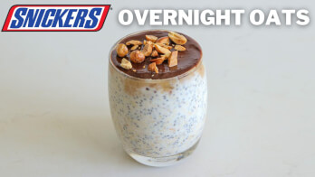 A glass filled with overnight oats topped with a layer of chocolate and sprinkled with chopped nuts. Text on the image reads "SNICKERS" in blue and red letters, and "OVERNIGHT OATS" in white letters above the glass.