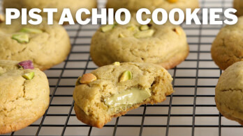 Close-up of several pistachio cookies cooling on a wire rack. One cookie in the foreground is partially eaten, revealing a green filling inside. Pistachio pieces are visible on top of the cookies. The text "PISTACHIO COOKIES" is displayed at the top of the image.