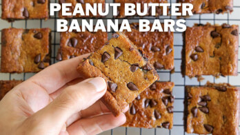 A hand is holding a square peanut butter banana bar with chocolate chips. More bars are arranged on a cooling rack in the background. The text "Peanut Butter Banana Bars" is displayed at the top.