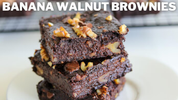 Three banana walnut brownies are stacked on a white plate. They are rich and dark in color, with visible walnut pieces embedded throughout. The text "BANANA WALNUT BROWNIES" is displayed at the top of the image. A cooling rack with more brownies is in the background.