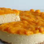 A cheesecake with a graham cracker crust, topped with a layer of bright yellow peach slices. A slice is being lifted from the whole cake, revealing the creamy, smooth interior of the cheesecake. The background is plain and white.