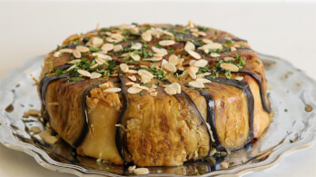 A round, savory dish is plated on a silver serving platter. The dish is wrapped in thin slices of roasted eggplant and topped with a garnish of chopped herbs and sliced almonds.