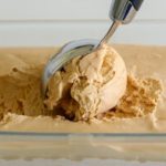 A metal ice cream scooper is scooping a creamy ball of coffee-flavored ice cream from a rectangular glass container filled with the same ice cream. The background is a light, neutral color.