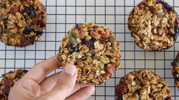 A hand holding a homemade granola cookie filled with oats, nuts, seeds, and dried fruit. Other similar cookies are cooling on a wire rack in the background.