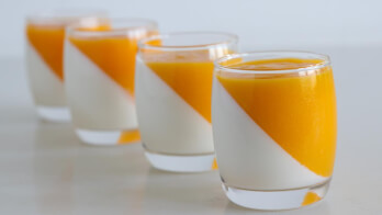 Four glasses filled with a layered dessert, featuring a diagonal split of white cream on the bottom and orange mango puree on the top, are arranged in a row on a white surface. The vibrant colors and smooth textures create an appealing presentation.