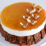 A round cake with a honeycomb-patterned topping and adorned with small, bee-shaped decorations made of fondant or marzipan. The sides of the cake are lined with honeycomb-like edible pieces, and the cake sits on a white plate. The background is plain, light-colored.