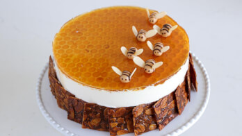 A round cake with a honeycomb-patterned topping and adorned with small, bee-shaped decorations made of fondant or marzipan. The sides of the cake are lined with honeycomb-like edible pieces, and the cake sits on a white plate. The background is plain, light-colored.