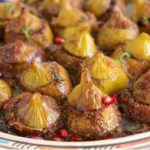 A colorful and ornate dish filled with roasted figs, garnished with a few pomegranate seeds and a hint of fresh herbs. The figs are caramelized and arranged neatly on a decorative plate, showcasing a rich glaze that highlights their texture and colors.