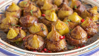 A colorful and ornate dish filled with roasted figs, garnished with a few pomegranate seeds and a hint of fresh herbs. The figs are caramelized and arranged neatly on a decorative plate, showcasing a rich glaze that highlights their texture and colors.