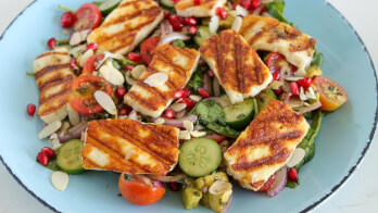 A fresh salad topped with grilled halloumi cheese, cherry tomatoes, cucumber slices, pomegranate seeds, almond flakes, and a mix of greens on a light blue plate. The grilled cheese has distinct grill marks and rests atop the vibrant vegetables.