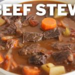A bowl of beef stew with chunks of beef, carrots, potatoes, and other vegetables in a thick broth. The words "BEEF STEW" are prominently displayed above the bowl.