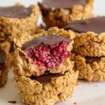 A close-up of stacked oat cups filled with a dark chocolate topping and a raspberry jam center. One of the cups has a bite taken out, revealing its vibrant raspberry filling. The oat cups are placed on a white surface.