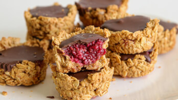 A close-up of stacked oat cups filled with a dark chocolate topping and a raspberry jam center. One of the cups has a bite taken out, revealing its vibrant raspberry filling. The oat cups are placed on a white surface.