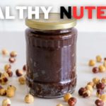 A jar of homemade Nutella sits on a white surface, surrounded by scattered hazelnuts. The text "HEALTHY NUTELLA" is prominently displayed above the jar, with "HEALTHY" in white and "NUTELLA" in red and black.