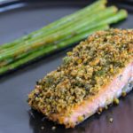 A serving of baked salmon fillet topped with a golden, crispy herb crust is plated alongside a bundle of tender asparagus spears. Both are presented on a dark, round plate.