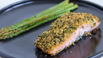 A serving of baked salmon fillet topped with a golden, crispy herb crust is plated alongside a bundle of tender asparagus spears. Both are presented on a dark, round plate.