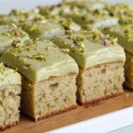 Sliced pieces of moist cake with a pale green frosting, topped with chopped pistachios. The cake rests on a wooden surface and appears to have a light, fluffy texture.