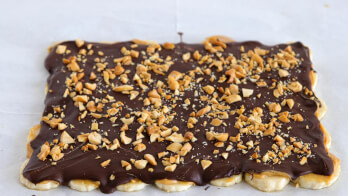 A rectangular dessert topped with smooth melted chocolate and sprinkled with chopped nuts on a white surface. The edges of the dessert show glimpses of a light, flaky pastry underneath the chocolate layer.