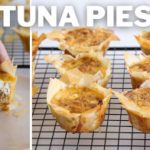 A close-up view of mini tuna pies on a cooling rack. On the left, a hand holds one tuna pie, revealing its filling. The pies have crispy, golden-brown crusts and rich, savory centers. The text "TUNA PIES" is prominently displayed at the top of the image.