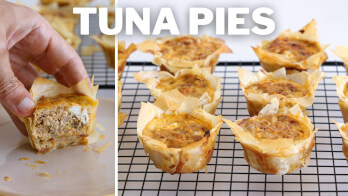 A close-up view of mini tuna pies on a cooling rack. On the left, a hand holds one tuna pie, revealing its filling. The pies have crispy, golden-brown crusts and rich, savory centers. The text "TUNA PIES" is prominently displayed at the top of the image.