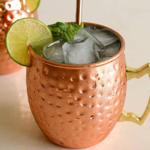 A close-up shot of a copper mug filled with a cocktail garnished with ice cubes, a lime wheel, and fresh mint leaves. A copper straw is placed in the drink, and another copper mug is slightly visible in the background.