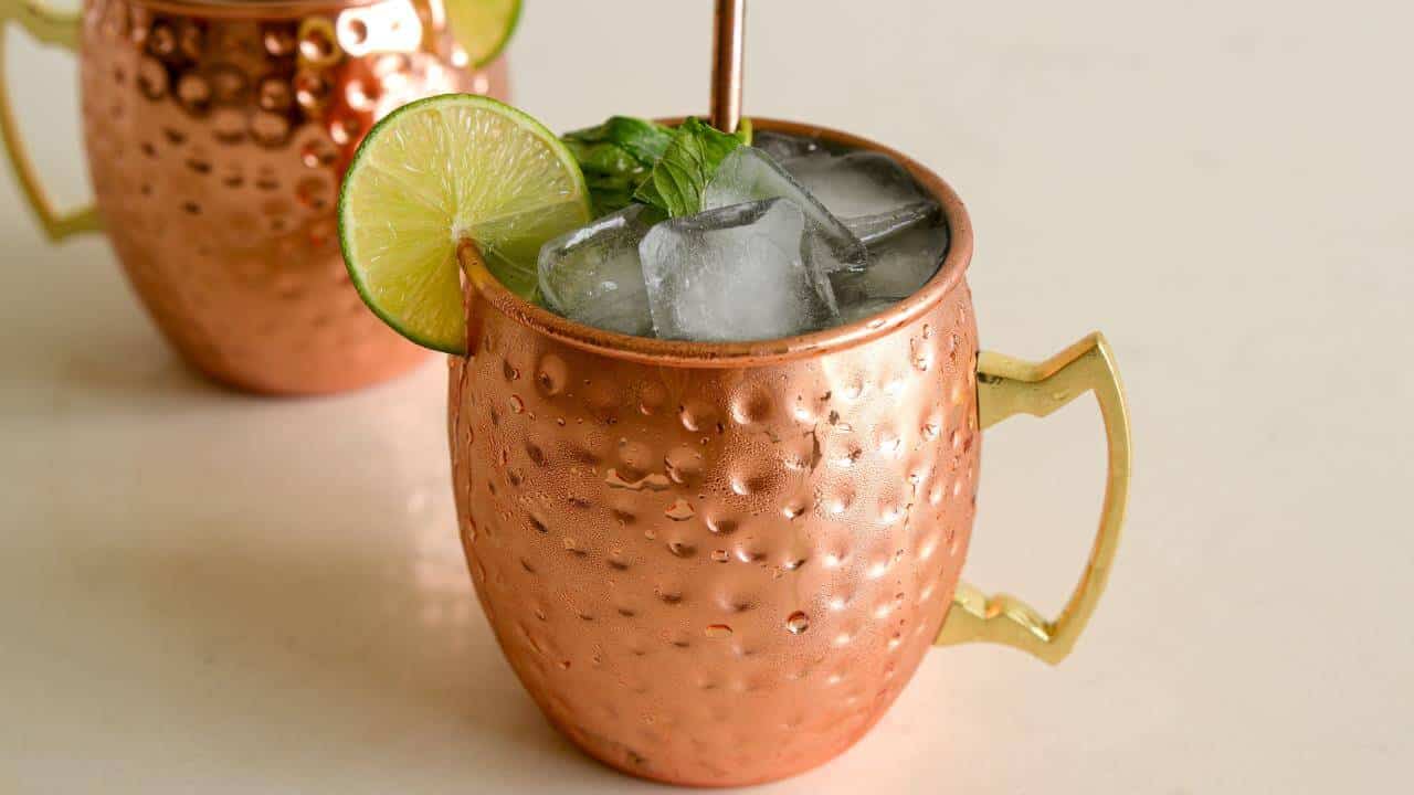 A close-up shot of a copper mug filled with a cocktail garnished with ice cubes, a lime wheel, and fresh mint leaves. A copper straw is placed in the drink, and another copper mug is slightly visible in the background.