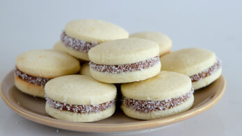 A beige plate with several alfajores, which are round sandwich cookies filled with dulce de leche and coated with a layer of desiccated coconut around the edges. The background is a plain, light-colored surface.