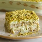 A square serving of creamy tiramisu topped with crushed pistachios on a beige plate. In the background, a glass dish holds more of the layered dessert with visible ladyfinger biscuits and cream layers.