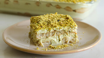 A square serving of creamy tiramisu topped with crushed pistachios on a beige plate. In the background, a glass dish holds more of the layered dessert with visible ladyfinger biscuits and cream layers.