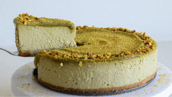 A rich green matcha cheesecake topped with crushed nuts on a white cake stand. A slice has been lifted from the cake, showcasing its creamy texture and a light brown crust.