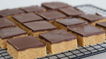 Close-up of multiple bars of chocolate-topped peanut butter fudge squares arranged in rows on a cooling rack. The bars have a thick layer of chocolate on top and a dense, golden-brown peanut butter layer underneath.