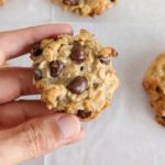 A person's hand holding a homemade oatmeal chocolate chip cookie. Several more cookies are placed on parchment paper in the background. The cookie appears chewy and full of oats and chocolate chips.