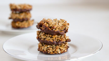 Three homemade sunflower seed and chocolate clusters are stacked on a white plate in the foreground, while additional clusters are visible on a similar plate in the background. The clusters appear to be bound together with a sweet, sticky coating.