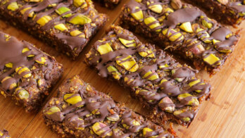 A close-up of several granola bars topped with chopped pistachios and drizzled with chocolate, arranged neatly on a wooden surface. The bars have a rich, textured appearance and the green pistachios contrast with the dark chocolate.