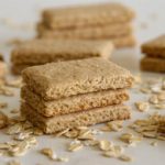 Stacked and scattered rectangular oat biscuits lie on a light surface, surrounded by a sprinkling of uncooked oats.