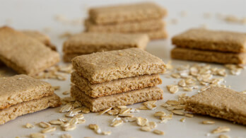 Stacked and scattered rectangular oat biscuits lie on a light surface, surrounded by a sprinkling of uncooked oats.