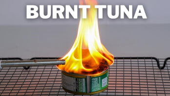 A can of tuna sits on a wire rack with intense flames shooting out from its top. The words "BURNT TUNA" are displayed prominently above the image.