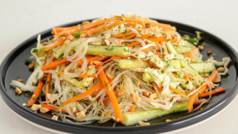 A black plate is filled with a colorful Asian noodle salad. The salad includes rice noodles, julienned carrots, cucumber slices, bean sprouts, and chopped peanuts, all topped with a light dressing and garnished with fresh herbs.