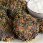 A plate featuring several golden-brown spinach fritters, served alongside a small bowl of creamy white dip. The fritters have a slightly crispy exterior and are placed on a white plate with blue floral patterns.