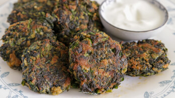 A plate featuring several golden-brown spinach fritters, served alongside a small bowl of creamy white dip. The fritters have a slightly crispy exterior and are placed on a white plate with blue floral patterns.