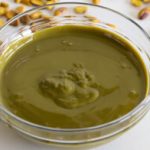 A clear glass bowl filled with creamy green pistachio paste sits on a white surface surrounded by scattered whole pistachios. The smooth texture of the paste contrasts with the crunchy nuts around it.