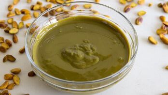 A clear glass bowl filled with creamy green pistachio paste sits on a white surface surrounded by scattered whole pistachios. The smooth texture of the paste contrasts with the crunchy nuts around it.