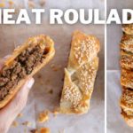 A close-up of a hand holding a slice of meat roulade, showing the savory meat filling inside flaky pastry. Next to it, a whole uncut meat roulade garnished with sesame seeds is placed on parchment paper. The text "MEAT ROULADE" is written at the top.